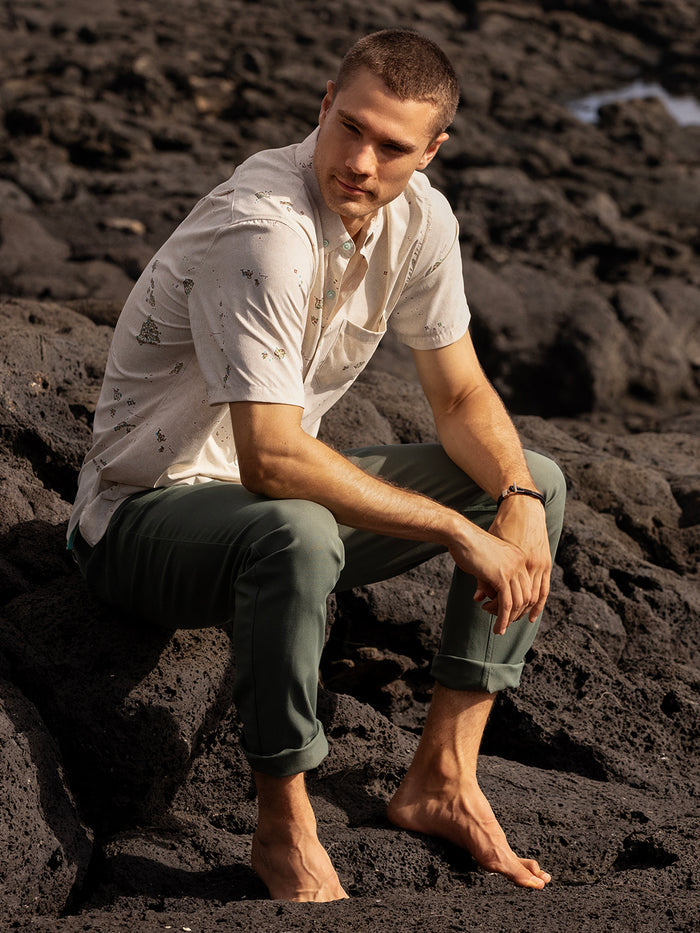 Beige tropical-print short-sleeve shirt and light rolled pants, seated outdoors on volcanic rocks.
