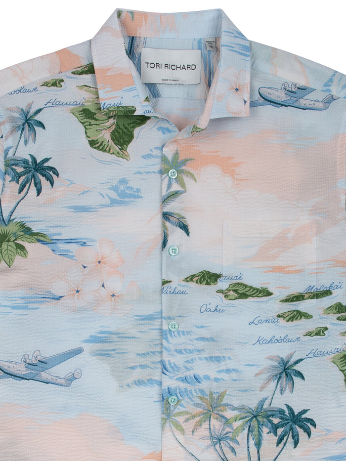 Light blue tropical-print button-up shirt with palms, planes, and island names, front view.