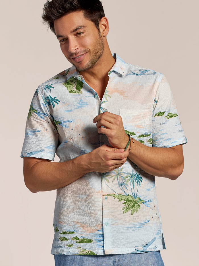 Light blue tropical palm and ocean, island and plane print cotton shirt man adjusting bracelet, front view.