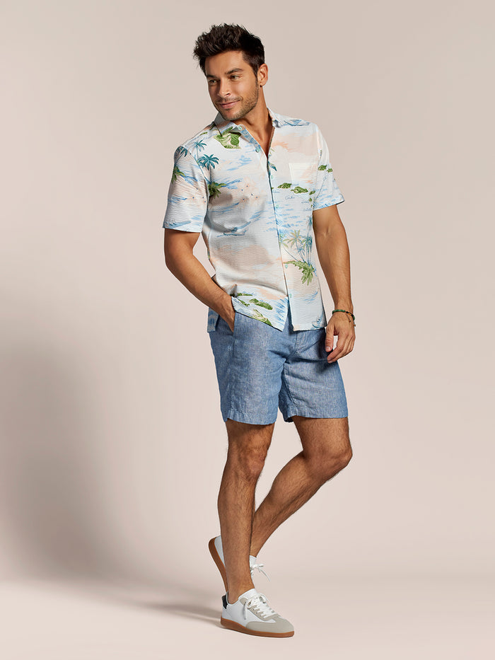 Light blue tropical palm and ocean, island and plane print cotton shirt, blue shorts, and white sneakers, full body of a man front view.