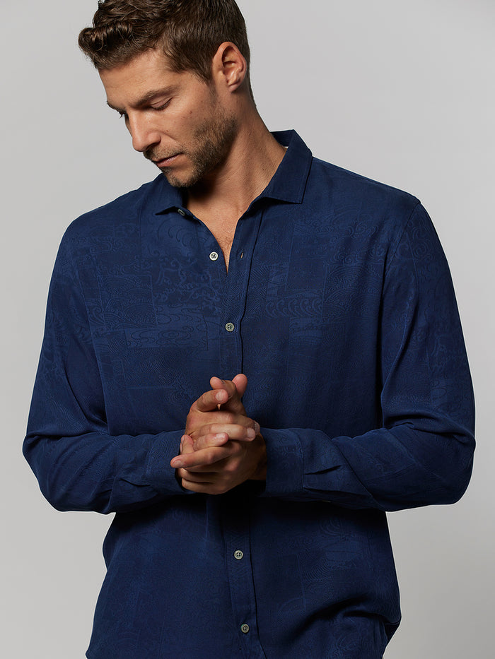 Man in blue Japanese wave-print long-sleeve shirt, looking down with hands clasped.