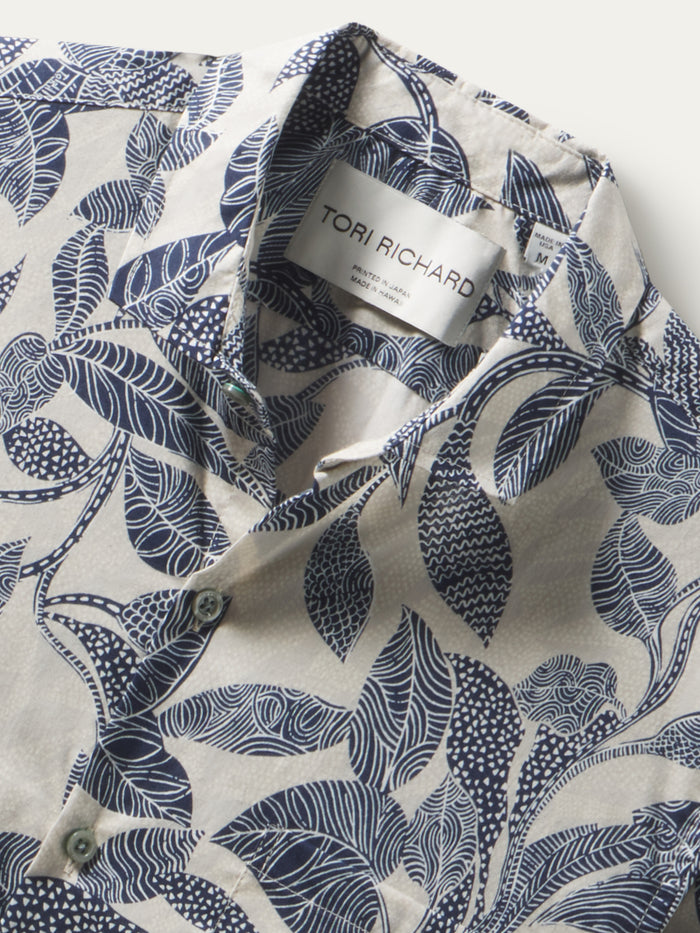 White short-sleeve button-up shirt with blue plumeria print, close-up fabric detail; leaf-print shirt, detailed fabric close-up view.