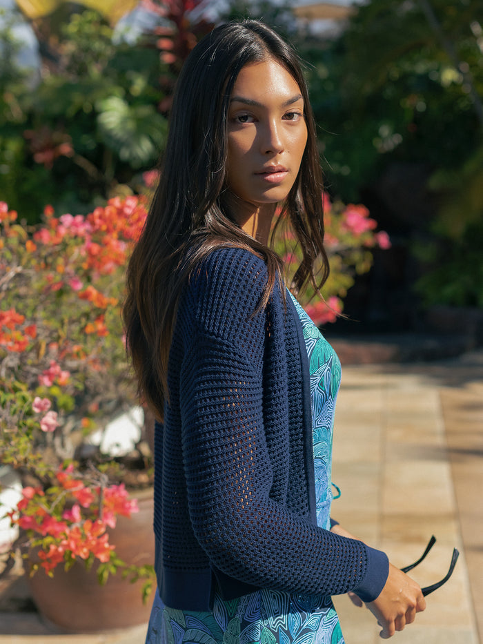 Woman outdoors in blue dress and light blue cardigan, holding sunglasses near flowers.