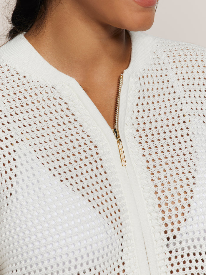 White pointelle knit cardigan with gold zipper, close-up of neck, shoulder, and partial face.