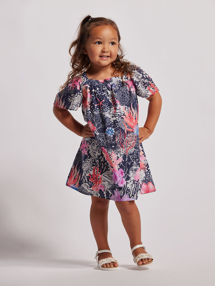 A young girl wearing a short-sleeved floral dress with a mix of pink, blue, and other colors.