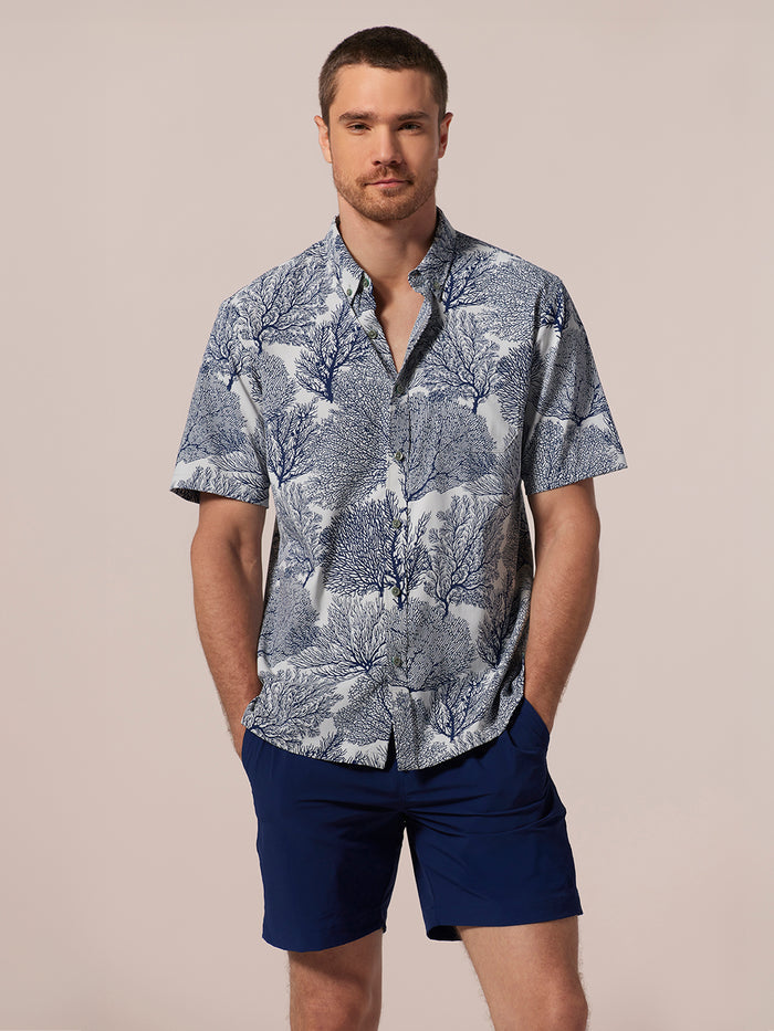 White short-sleeve shirt with navy coral pattern/print, button-down short-sleeve shirt and dark blue shorts, model standing hands in pockets, front view.