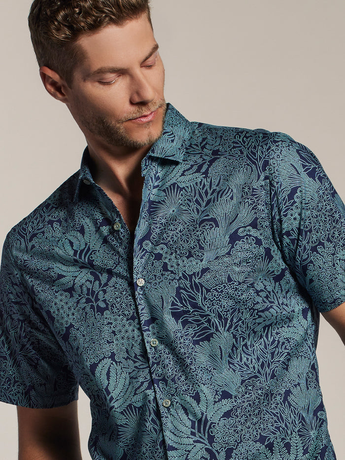 Man in blue patterned short-sleeve shirt, looking down, front view, neutral background.
