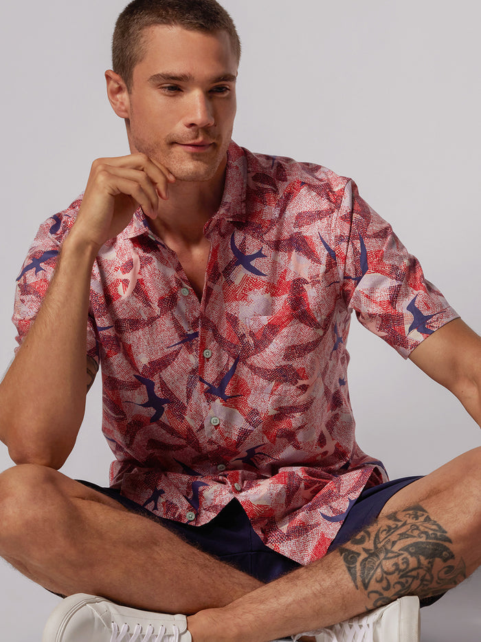 Red logo-print short-sleeve shirt, navy shorts, and white sneakers, seated cross-legged.