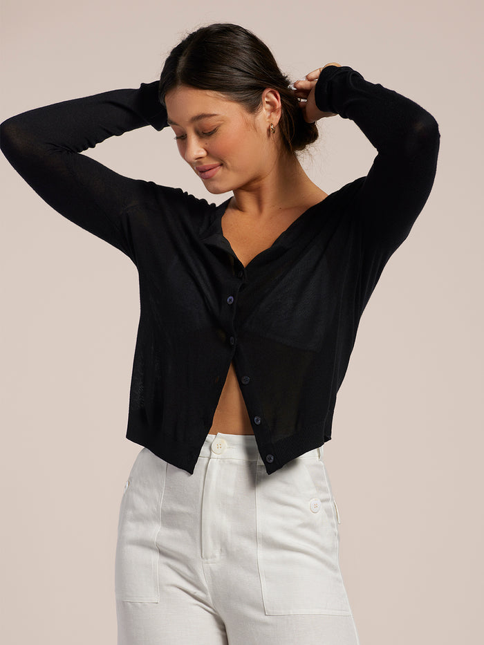 Black knit cardigan, white pants, arms raised, smiling, front view.