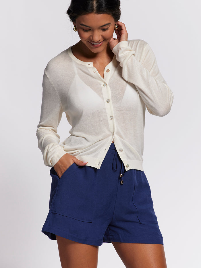 Woman in cream Featherweight Knits Nina cardigan and blue shorts, smiling with hand on face, front view.