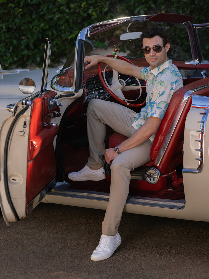 A man wearing Beige short-sleeve shirt with blue and green leaf print, white sneakers, in vintage convertible, front and side views.