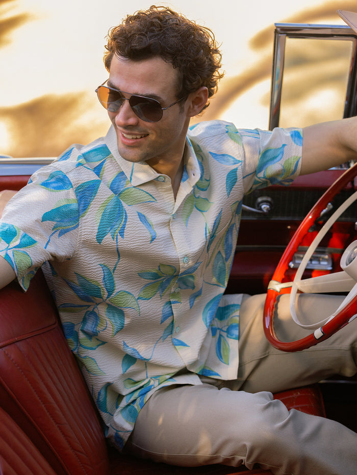 Beige short-sleeve shirt with blue and green leaf print. a man seated in red vintage car, front view.