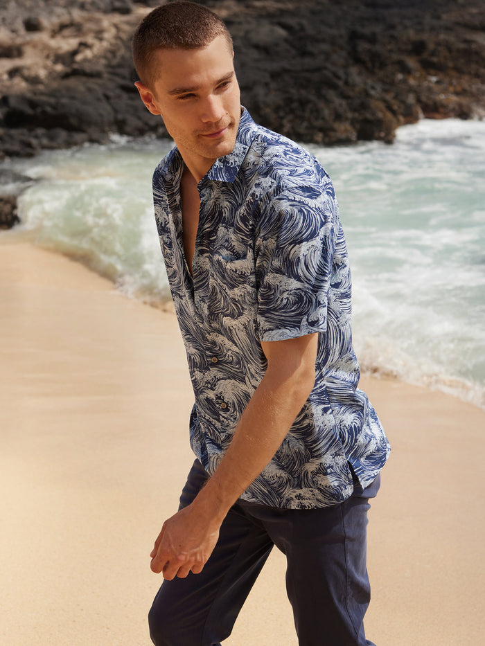 A man wearing the Gnarly blue and white patterned Cotton Lawn short-sleeve shirt and blue pants strolls along a sandy beach near the ocean, rocky cliffs behind him, highlighting timeless Hawaiian style.