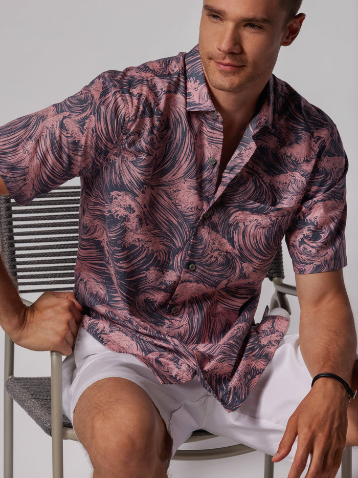 A man sits on a chair wearing the Gnarly short-sleeve cotton lawn shirt, featuring a pink and dark blue wavy pattern inspired by Japanese art, paired with white shorts. He rests one hand on the chair and looks to the side, smiling slightly.