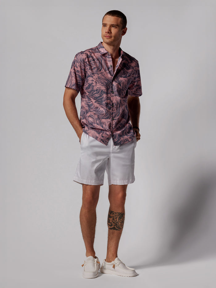 A man stands against a light gray background wearing the Gnarly short-sleeve Cotton Lawn shirt with a pink and purple pattern inspired by Japanese art, paired with white shorts and sneakers. His hands are in his pockets and he has a tattoo on his left calf.