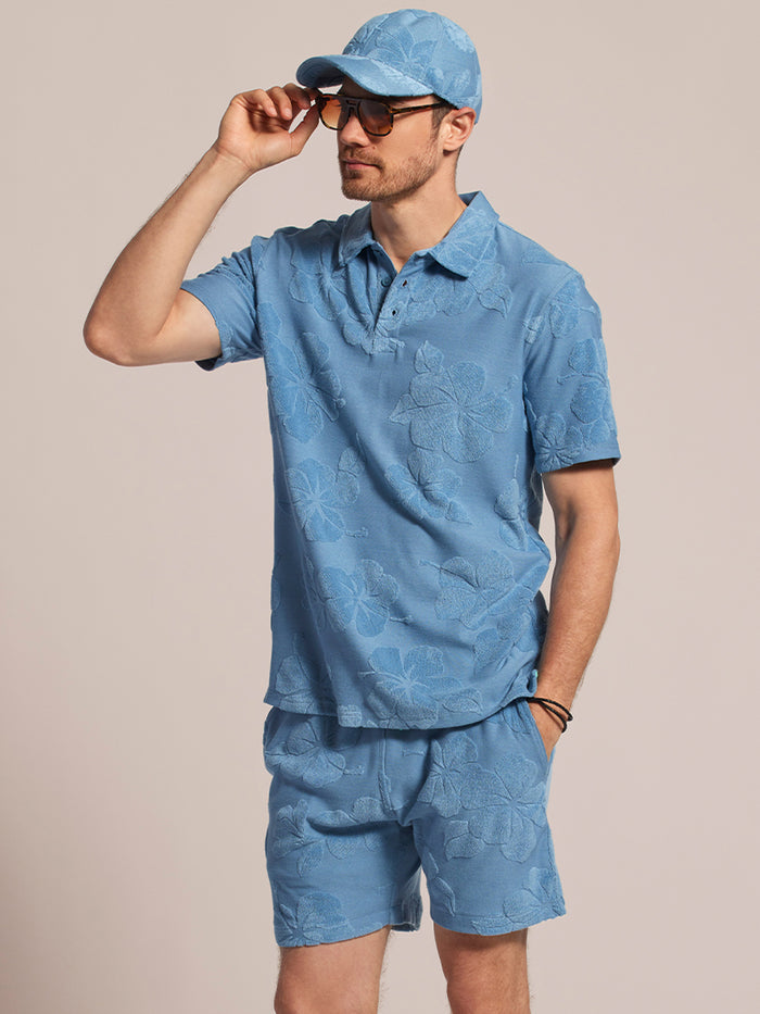 Green polo and navy hibiscus-print shorts, front view, model adjusting sunglasses.