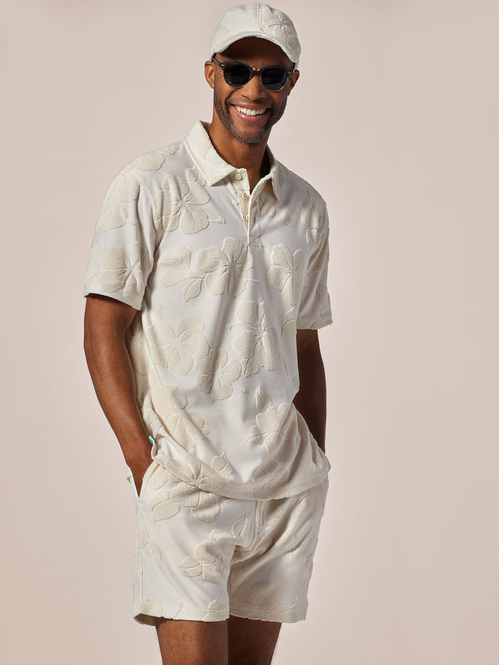 Beige hibiscus-print short-sleeve polo and shorts set, front view with cap and sunglasses.