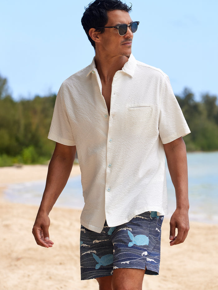 Blue print swim shorts, front view, worn by man in white shirt and sunglasses on beach.