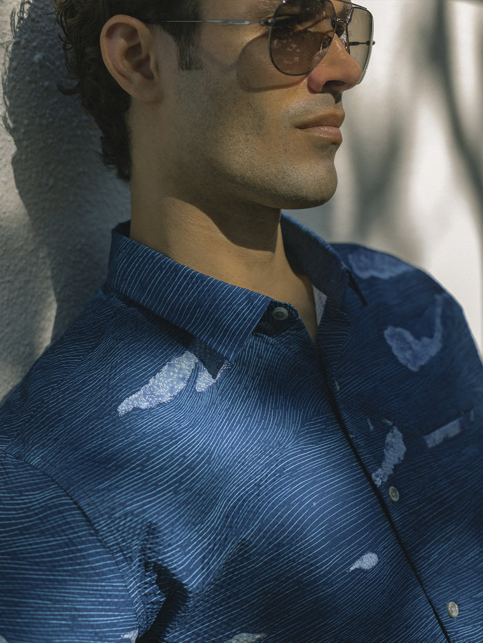 Blue solid cooling short-sleeve shirt, man leans against sunlit wall, side view with sunglasses.