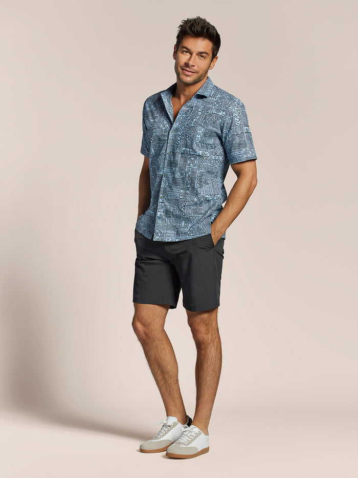 Blue short-sleeve button-up with black abstract Hawaiian geometric pattern, black shorts, and white sneakers, front view of a man.