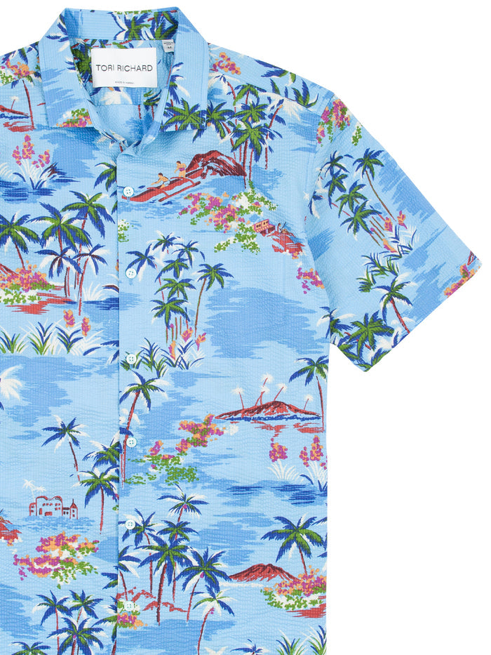Blue Hawaiian shirt with palm tree, ocean, and canoe print/pattern, front view.