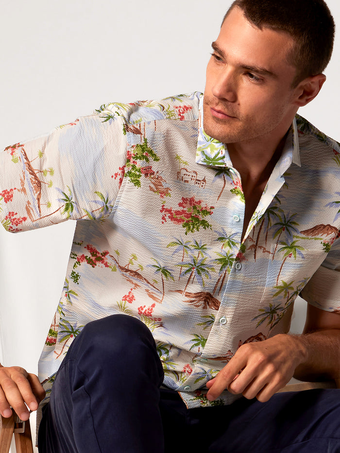 Cream tropical-print short-sleeve shirt, navy pants, seated man, side view.