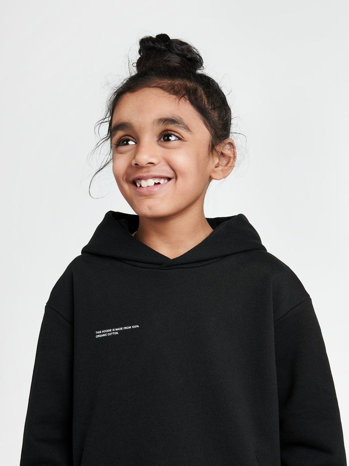 Kids' 365 Midweight black hoodie with white logo, 100% organic cotton, boxy oversized fit worn by smiling girl
