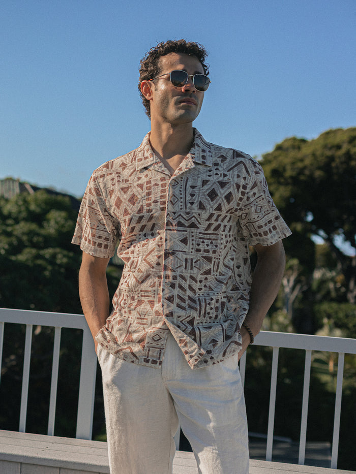 Beige short-sleeve button-up shirt with brown geometric tribal pattern palm-print short-sleeve shirt, worn by man in sunglasses, front view.