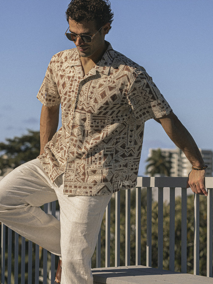 Beige short-sleeve button-up shirt with brown geometric tribal pattern, worn by man leaning on railing outdoors.