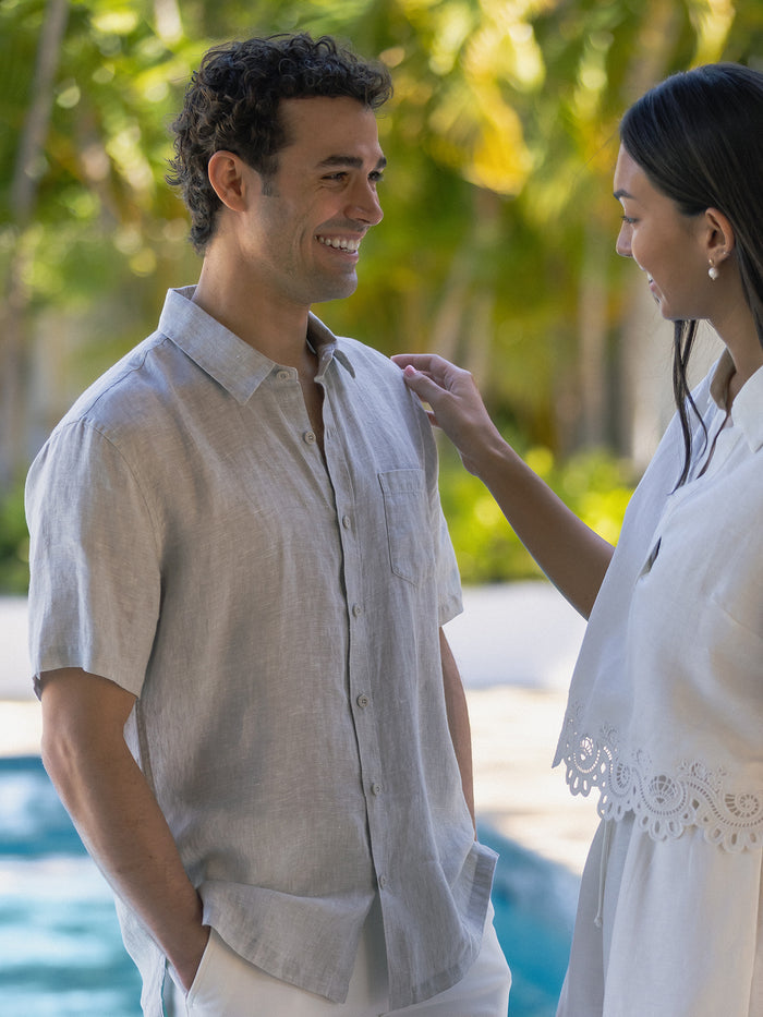 Kona Light blue linen short-sleeve shirt, poolside with palms. White sleeveless sundress, side view by tropical resort pool.