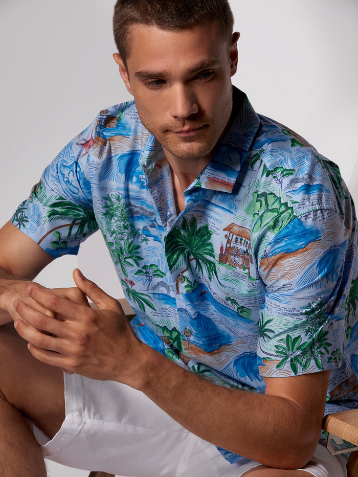 Blue tropical print short-sleeve shirt and white shorts, seated with journal in hand.