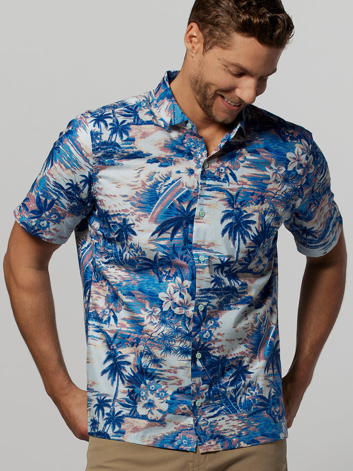 Blue palm tree print short-sleeve shirt, front view; model looking down and smiling.