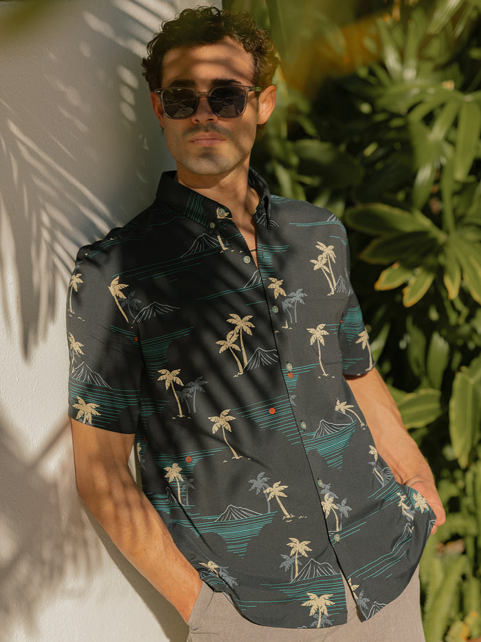 Black short-sleeve shirt with beige palm and teal mountain print, palm print, outdoor front view with sunglasses on a model outdoor.