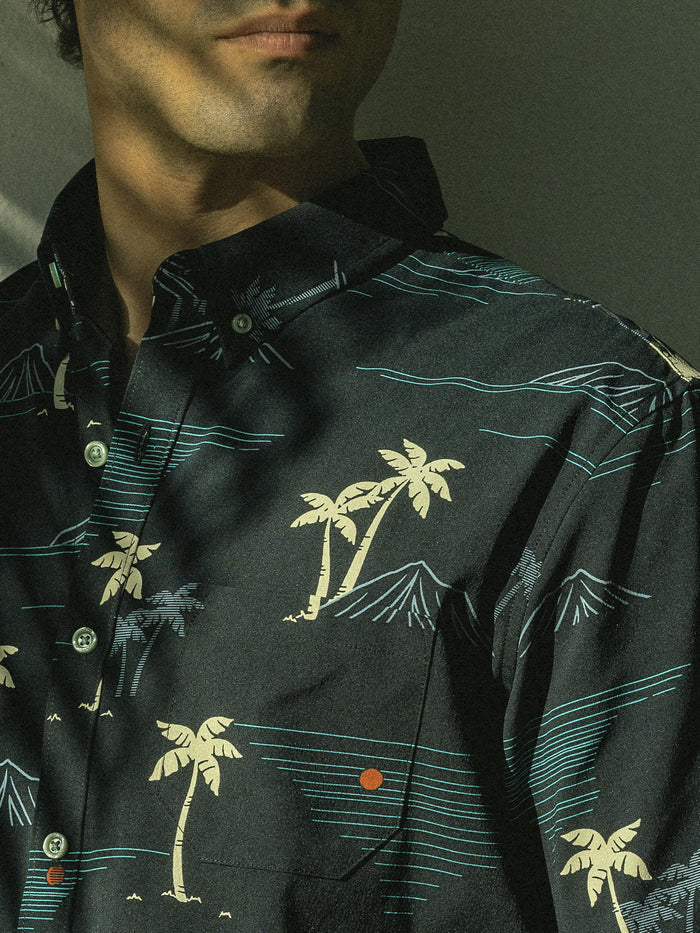 Black short-sleeve shirt with beige palm and teal mountain print, palm print, sunlit front view.