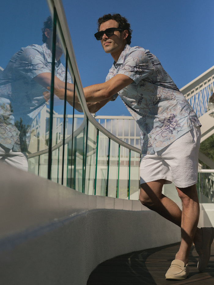 Man in octopus-print short-sleeve shirt and white shorts leans on glass railing.