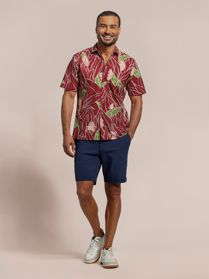 Red tropical floral-print short-sleeve cotton shirt with green leaves and pink flower print and navy shorts, front view of a man.
