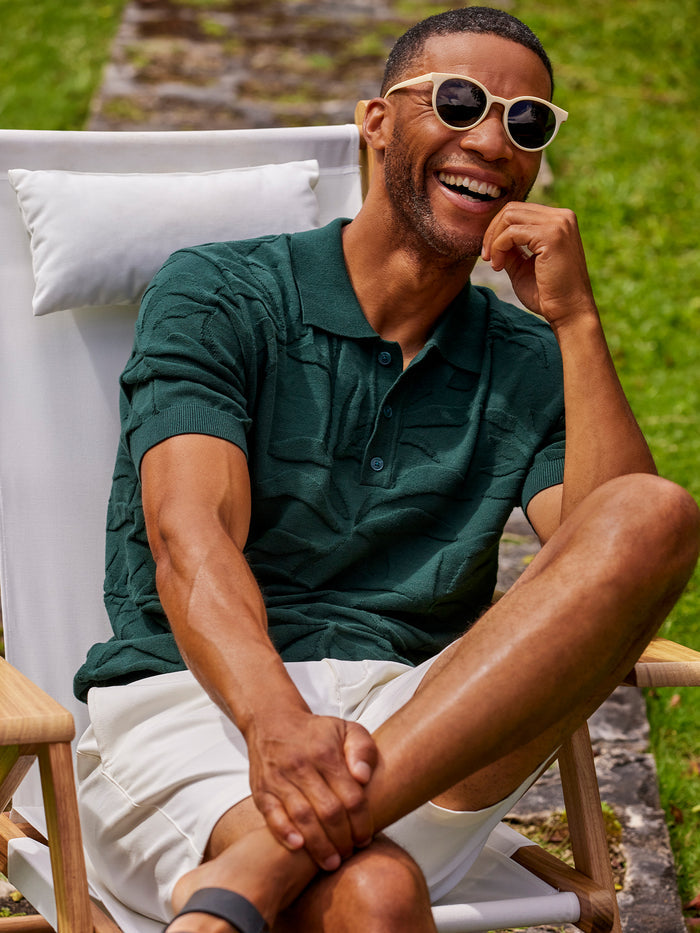 Dark green palm-print sweater polo shirt and sunglasses seated or lounging outdoors, front view.