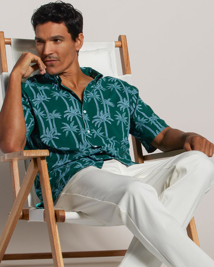 Green short-sleeve shirt with blue palm print and white pants, man seated outdoors, front view.