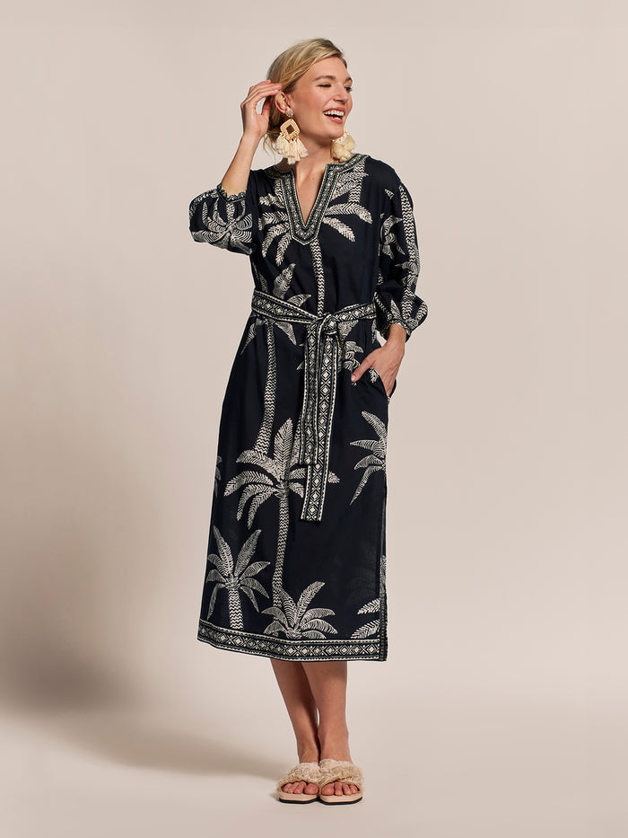 Palm-print belted caftan dress with large earrings and sandals, hand near face, front view.
