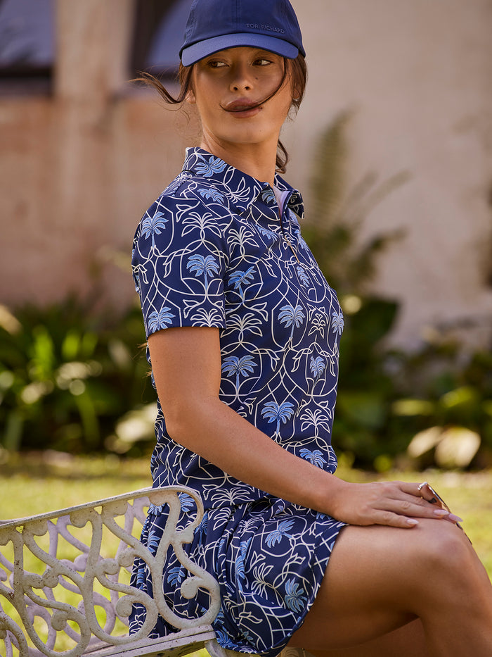 Blue palm-print short-sleeve polo shirt, outdoor seated among greenery.