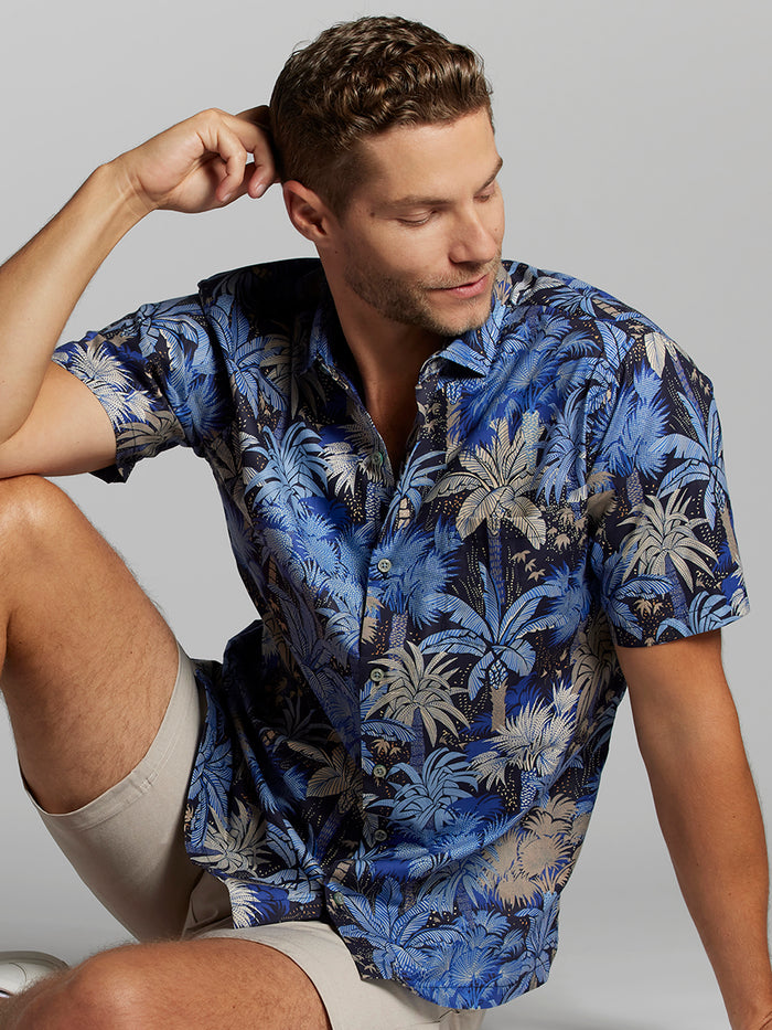 Blue, beige, and gray palm-print short-sleeve shirt with beige shorts, sitting, side view.