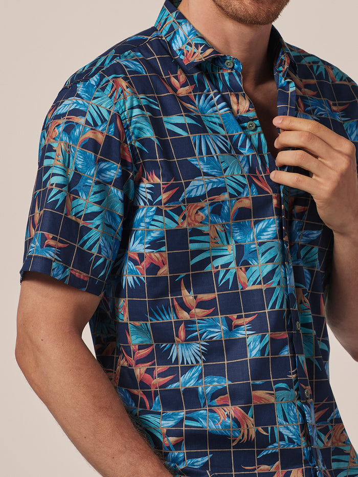 Blue and orange tropical leaf floral and grid-patterned short-sleeve shirt, front view, man adjusting collar.