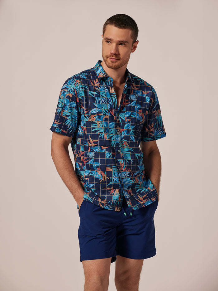 Blue and orange tropical leaf -print short-sleeve shirt and blue shorts, front view, hands in pockets.