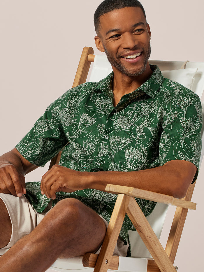 Green floral-print short-sleeve shirt and beige shorts, seated front view.