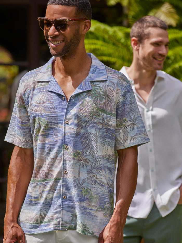 Blue and green tropical-print short-sleeve shirt with green pants, White cabana-print short-sleeve shirt, sunglasses, outdoor view.