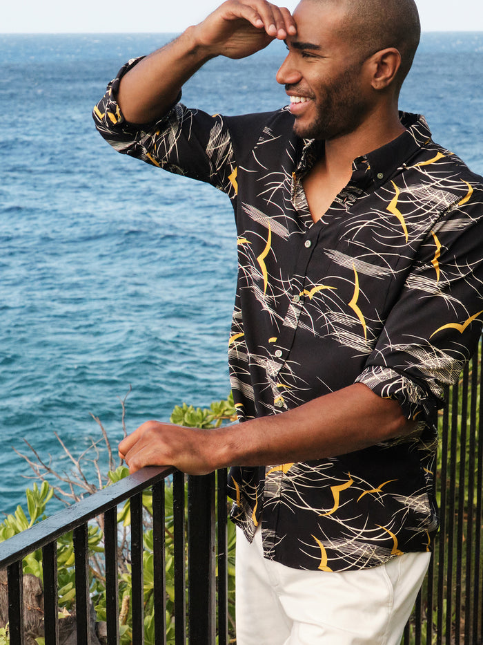 Black long-sleeve shirt with abstract wave print, and golden seagull print relaxed fit, shown outdoors leaning on railing with ocean backdrop.