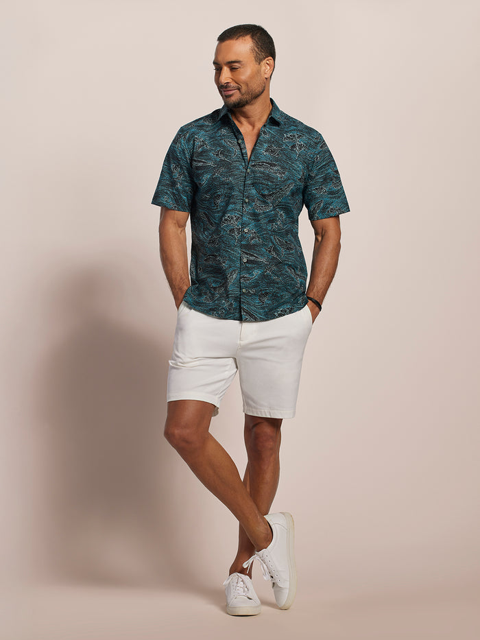 Blue short-sleeve shirt with black and gray abstract ocean and black whale print, front view of a man with white shorts and sneakers.