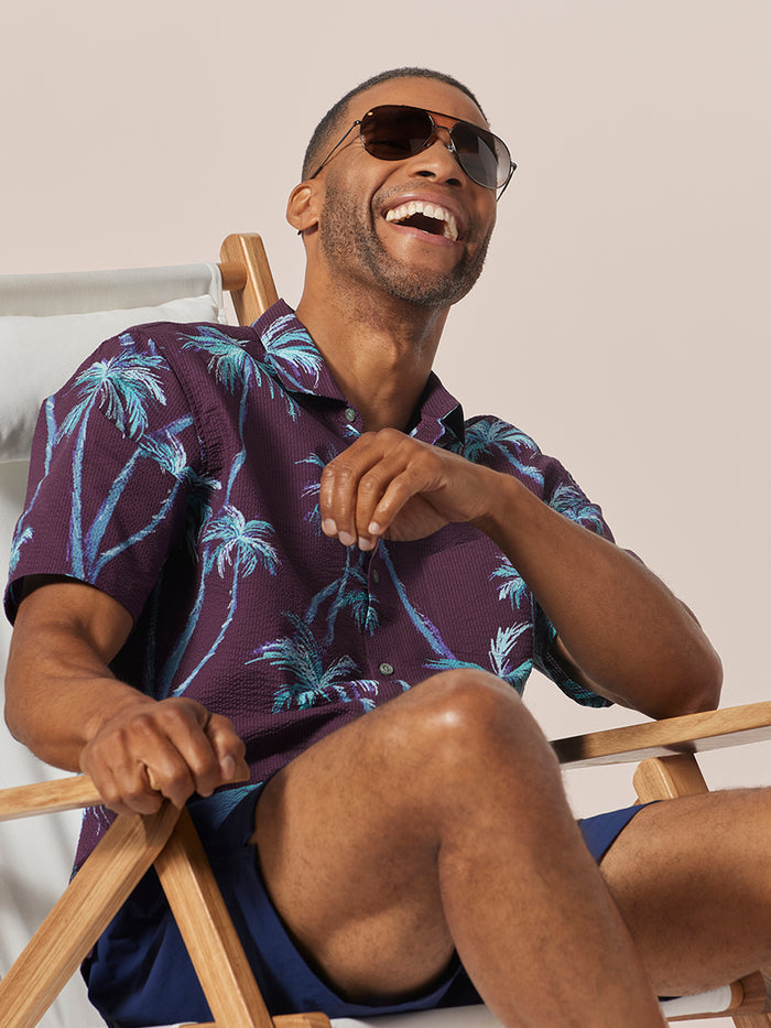 Blue and purple palm-print short-sleeve shirt, man seated on wooden chair, front view.