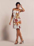 Woman in light floral-print midi dress and strappy heels smiles, touching hair in studio setting.