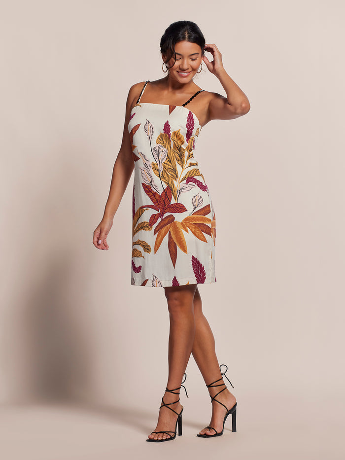 Woman in light floral-print midi dress and strappy heels smiles, touching hair in studio setting.